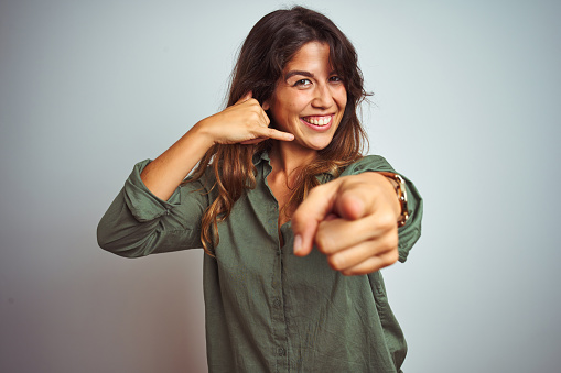 Young beautiful woman wearing green shirt standing over grey isolated background smiling doing talking on the telephone gesture and pointing to you. Call me.