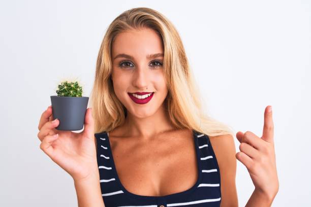 Young beautiful woman holding small cactus pot standing over isolated white background surprised with an idea or question pointing finger with happy face, number one