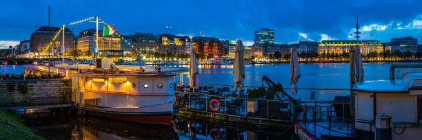 Panoramic view across the blue waters of Binnen-Alster lake and its famous fountain to the warm lights of the stores, hotels and restaurants on Jungfernstieg in the heart of Hamburg, Germany’s vibrant second city.