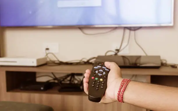 Close up of Human Hand holding a Remote control and pointed towards a TV screen for changing or zapping channels. Modern technology and lifestyle concept.