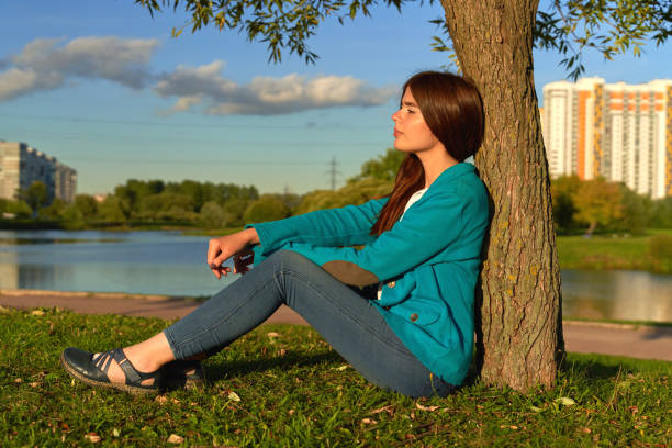 Summer shot of young attractive woman with beautiful long brunette hair, sitting under tree, resting near the pond with pensive thoughtful expression against green and urban background of big city.