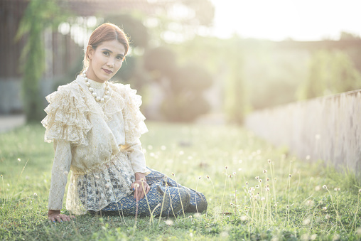 Asian woman bride in thai traditional costume sit on grass at outdoor in a morning surrounding by golden sunlight and grass flower.