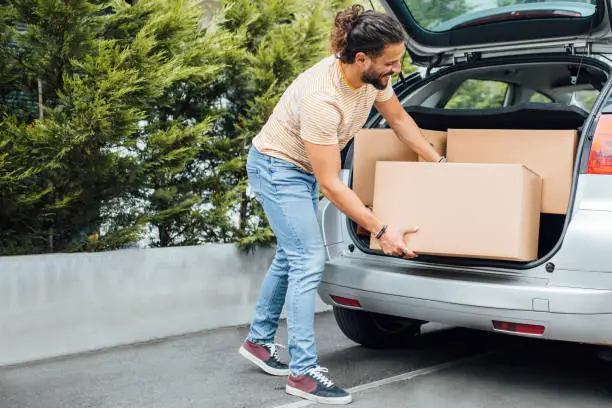 Young millennial man with long hair and a beard, putting boxes in the back of the car, moving