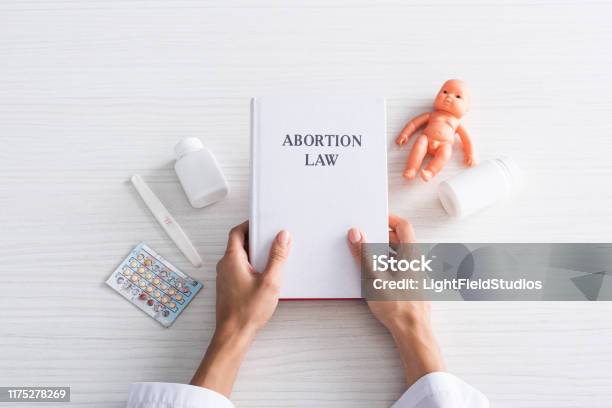 Top View Of Woman Holding Book With Abortion Lettering Near Baby Doll And Abortion Pills Stock Photo - Download Image Now