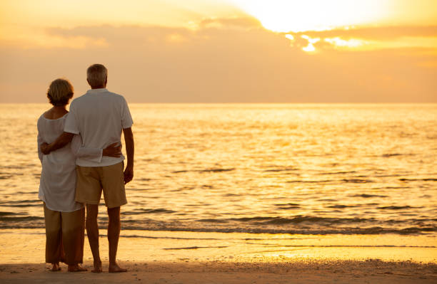 senior man and woman couple embracing at sunset or sunrise on a deserted tropical beach - men beach back rear view imagens e fotografias de stock