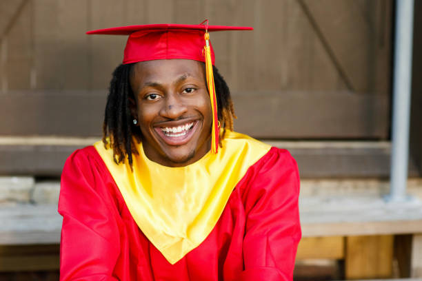 sonriendo feliz afroamericano adolescente adolescente hombre hombre fuera contra una pared marrón en su vestido de graduación rojo y dorado - poses para fotos de graduación fotografías e imágenes de stock