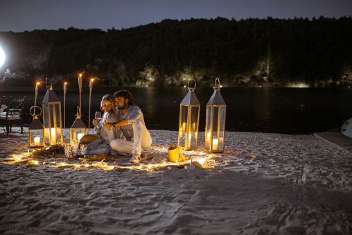 Couple having a romantic dinner at the beautifully decorated place illuminated with different lights the sandy beach at dusk. Wide view with copy space