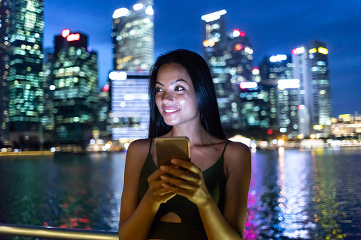 Beautiful young woman with long black hair using smart phone at night, in the city.