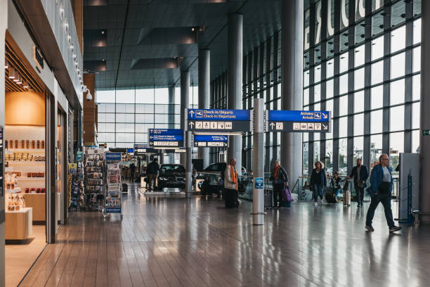 Travelers walk through a modern airport terminal with large windows, blue overhead signs, and nearby shops.