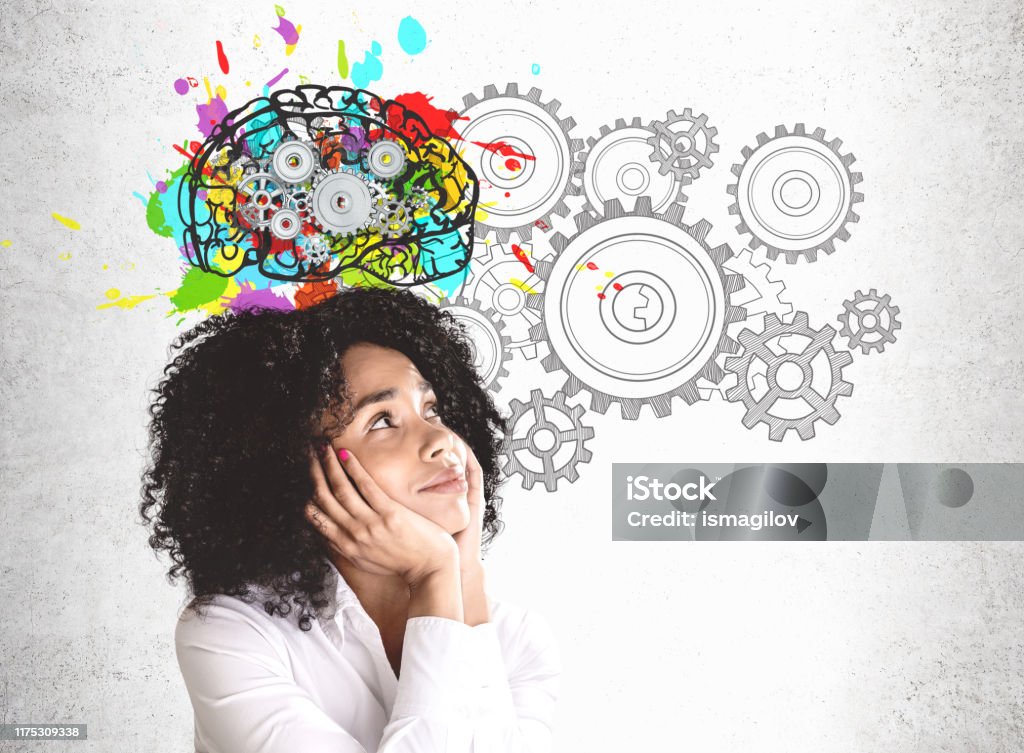Thoughtful young African woman brainstorming Smiling young African American woman in white shirt looking at colorful brain sketch with gears drawn on concrete wall. Concept of brainstorming Contemplation Stock Photo