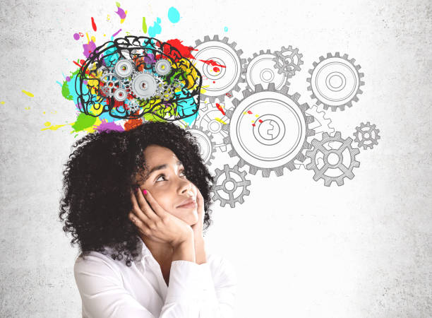 Thoughtful young African woman brainstorming Smiling young African American woman in white shirt looking at colorful brain sketch with gears drawn on concrete wall. Concept of brainstorming creativity stock pictures, royalty-free photos & images