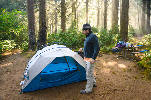 This is a photograph of an American man in his 30s standing by a tent at a state park campsite in the woods along the Oregon coast. Rays of sunlight shine through the trees in the background.