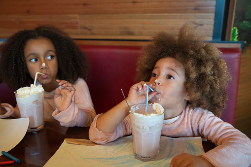 Ethnic kid sisters drinking smoothie ice cream at restaurant having fun