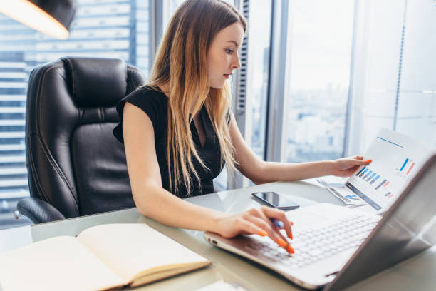 책상에 앉아 사무실에서 일하는 여성 이사는 노트북을 사용하여 다이어그램과 차트를 들고 비즈니스 통계를 분석합니다. - diagram business writing women 뉴스 사진 이미지