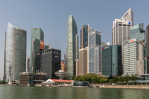 Singapore - March 21, 2019: On Marina waters. Skyscrapers of Financial District surround Clifford Square with pier and restaurant, including Customs House tower. All under blue sky and behind greenish water.