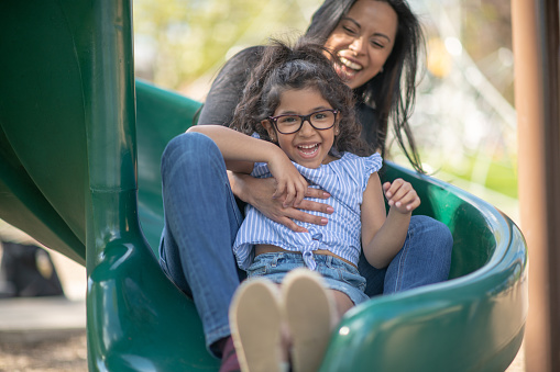 A young hispanic mother and daughter are playing together at the park. They are sliding down a slide and laughing with excitement. They are wearing casual clothes and the young child is wearing reading glasses.