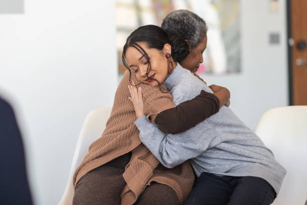 Supportive women hug while attending a group therapy session A young mixed-race woman hugs a mature adult black woman. They are sitting next to each other in a medical clinic. The two women are attending a group therapy session. They are showing support and kindness. mercy hug stock pictures, royalty-free photos & images