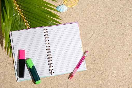 Travellers notebook with markers and pen on sand with palm tree leaf background, top view. Planning and ideas vacation concept. Flat lay style with copy space