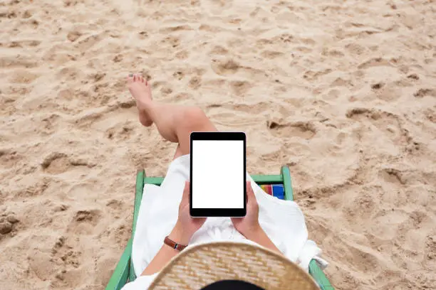 Photo of a woman holding and using a black tablet pc with blank desktop screen while lying down on a beach chair