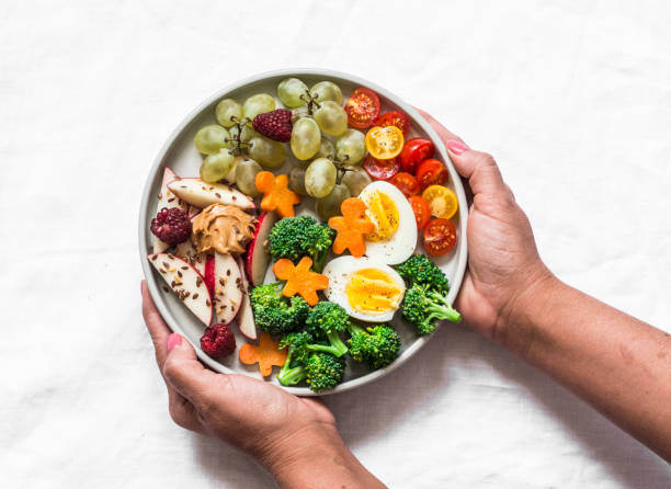 istock Female hands holding a large snack plate. Boiled egg, broccoli, carrots, apples with peanut butter, grapes - delicious diet breakfast, snack on a light background, top view 1175395604