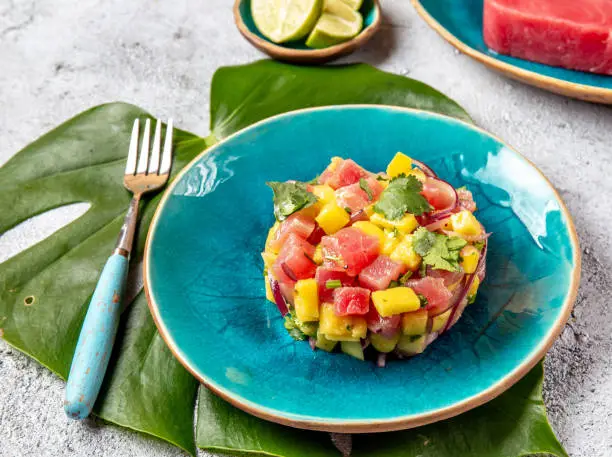 Tropical style gourmet tuna mango salad tartar with cilantro and purple onion. On blue plate, tropical leaves.