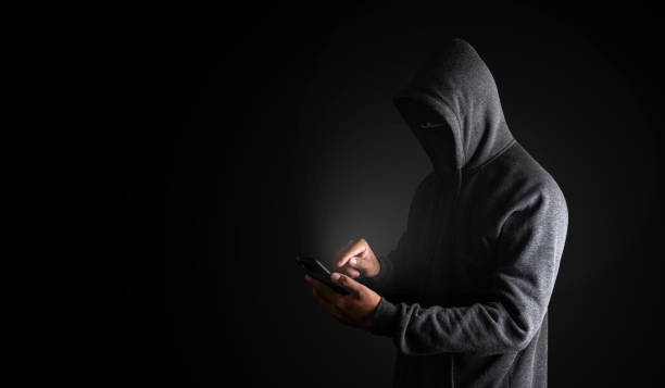 istock Hacker Using Smartphone. Men in black clothes with hidden face looks at smartphone screen on black background with copy space. with clipping path. 1175397194