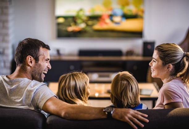 genitori felici che parlano mentre guardano la tv con i loro figli a casa. - family television watching watching tv foto e immagini stock