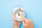Man hand holding white alarm clock. Finger pointing to arrow of twelve o'clock. Light pastel blue background. Time change concept. Closeup. Top view.