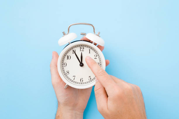 Man hand holding white alarm clock. Finger pointing to arrow of twelve o'clock. Light pastel blue background. Time change concept. Closeup. Top view. Man hand holding white alarm clock. Finger pointing to arrow of twelve o'clock. Light pastel blue background. Time change concept. Closeup. Top view. time zone stock pictures, royalty-free photos & images