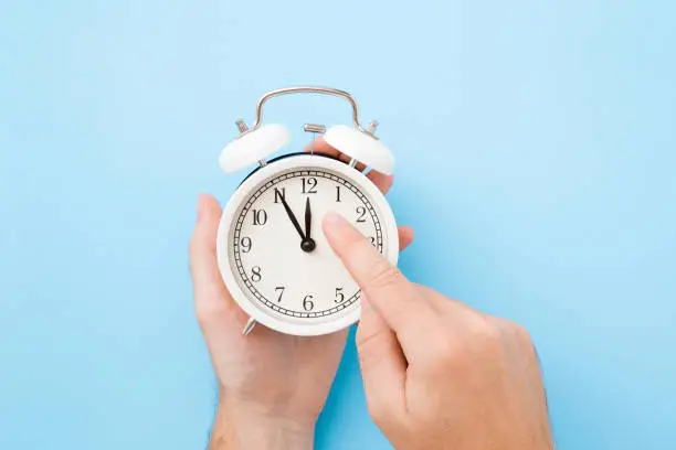 Photo of Man hand holding white alarm clock. Finger pointing to arrow of twelve o'clock. Light pastel blue background. Time change concept. Closeup. Top view.