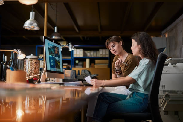 due creativi che condividono un computer e fanno un lavoro cartaceo. - piccolo ufficio foto e immagini stock