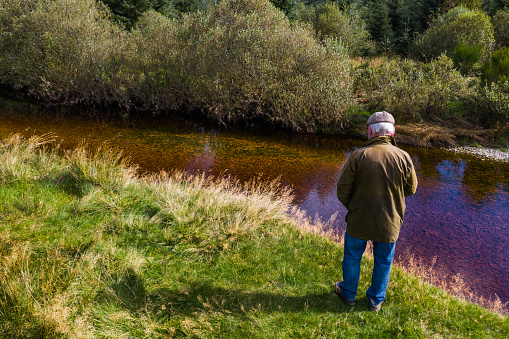 Senior man wearing a waxed jacket walking next to a river in rural south west Scotland