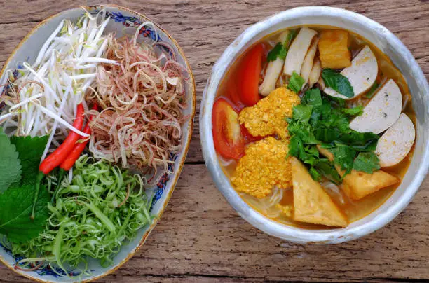 Top view bowl of homemade vegan rieu noodle soup or vegetarian crab paste vermicelli soup; a traditional Vietnamese dish; plate of vegetables; herbs; ready to eat on wooden background