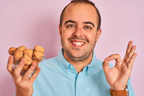 istock Young man holding bowl with walnuts standing over isolated pink background with happy face smiling doing ok sign with hand on eye looking through fingers 1175439809