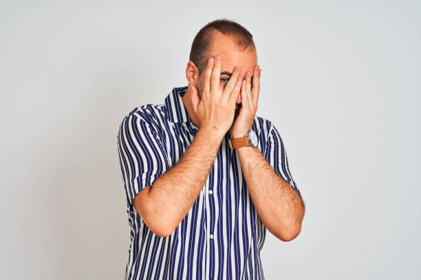 istock Young man wearing blue striped shirt standing over isolated white background with sad expression covering face with hands while crying. Depression concept. 1175440128