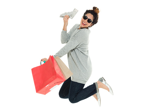 Full length / one person / side view / profile view of 30-39 years old adult beautiful puerto rican ethnicity / latin american and hispanic ethnicity female / young women / one young woman only shopaholic jumping / mid-air / standing in front of white background wearing sweater / jeans / sunglasses who is smiling / happy / cheerful / joy / excited / cool attitude who is shopping / buying and holding currency / us currency / shopping bag / bag / investment / savings / us paper currency / wealth / american one hundred dollar bill / sale / retail