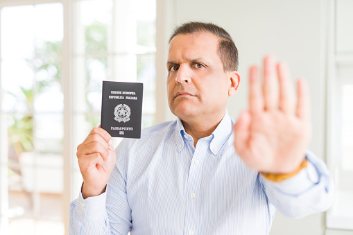 Middle age man holding holding passport of Italy with open hand doing stop sign with serious and confident expression, defense gesture
