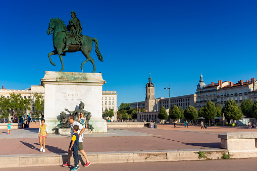Overview of the Place Bellecour, It is one of the largest open squares in Europe. In the middle is an equestrian statue of King Louis XIV by François-Frédéric Lemot. Tourists are walking across the square with a young family in the foreground. A typical summer day in Lyon, France