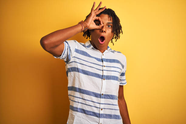 istock Afro american man with dreadlocks wearing striped shirt over isolated yellow background doing ok gesture shocked with surprised face, eye looking through fingers. Unbelieving expression. 1175449671