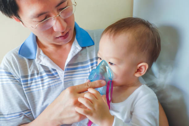 Asian father helping his toddler son with inhalation therapy by the mask of inhaler Asian father helping his toddler son with inhalation therapy by the mask of inhaler. Sick little kid with respiratory problem with oxygen mask breathes through nebulizer at doctor clinic / hospital pediatric nebulizer mask stock pictures, royalty-free photos & images