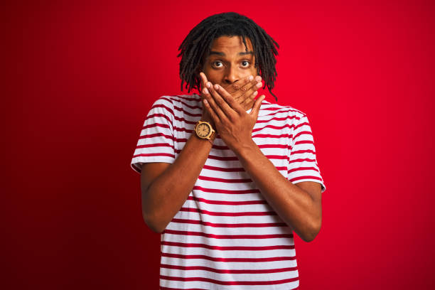istock Young afro man with dreadlocks wearing striped t-shirt standing over isolated red background shocked covering mouth with hands for mistake. Secret concept. 1175452100