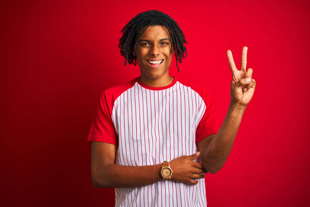 istock Afro man with dreadlocks wearing striped t-shirt standing over isolated red background smiling with happy face winking at the camera doing victory sign. Number two. 1175452386