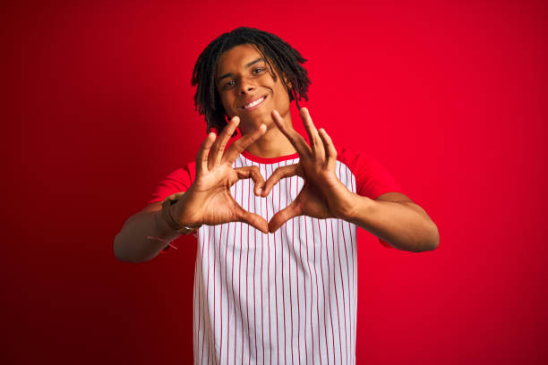 istock Afro man with dreadlocks wearing striped t-shirt standing over isolated red background smiling in love showing heart symbol and shape with hands. Romantic concept. 1175452405