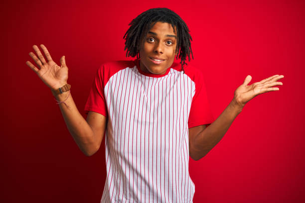 istock Afro man with dreadlocks wearing striped t-shirt standing over isolated red background clueless and confused expression with arms and hands raised. Doubt concept. 1175452939