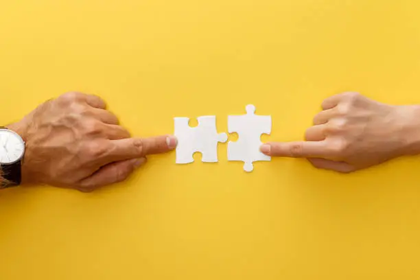 Photo of cropped view of woman and man matching pieces of white jigsaw puzzle on yellow background