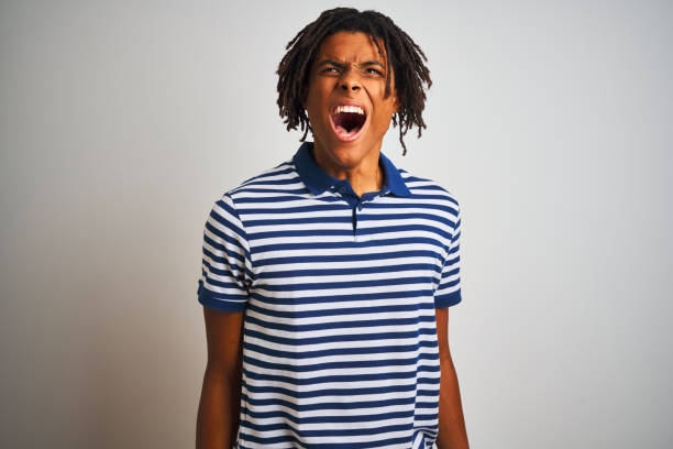 istock Afro man with dreadlocks wearing striped blue polo standing over isolated white background angry and mad screaming frustrated and furious, shouting with anger. Rage and aggressive concept. 1175454706