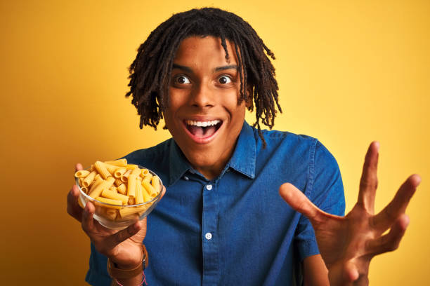 istock Afro american man with dreadlocks holding pasta macaroni over isolated yellow background very happy and excited, winner expression celebrating victory screaming with big smile and raised hands 1175456513