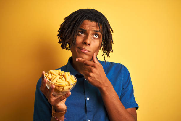 istock Afro american man with dreadlocks holding pasta macaroni over isolated yellow background serious face thinking about question, very confused idea 1175456534