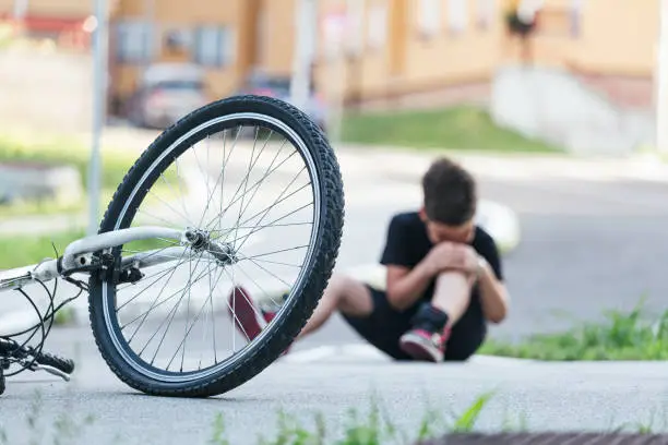 Kid hurts his leg after falling off his bicycle. Child is learning to ride a bike. Boy in the street ground with a knee injury screaming after falling off to his bicycle.