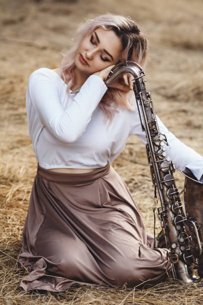 porträt einer schönen jungen frau sitzt auf dem trockenen gras leanher kopf auf saxophon, romantische blonde mädchen enge augen und entspannend - close up musical instrument saxophone jazz stock-fotos und bilder